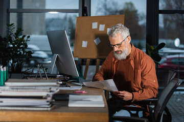 grey haired businessman working with papers while sitting at workplace near computer monitor in office