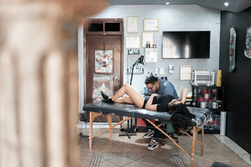 A tattoo artist is tattooing a snake on a woman's leg in his work studio