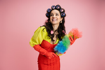 cheerful housewife in yellow blouse and hair curlers holding colorful dust brush and looking away isolated on pink