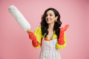 smiling housewife in striped apron and red rubber gloves holding white dust brush and waving hand isolated on pink