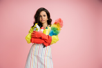 Obraz na płótnie Canvas frowning housewife in striped apron and red rubber gloves holding pile of cleaning supplies isolated on pink