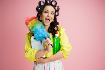 astonished housewife in hair curlers holding detergents and bright feather duster isolated on pink