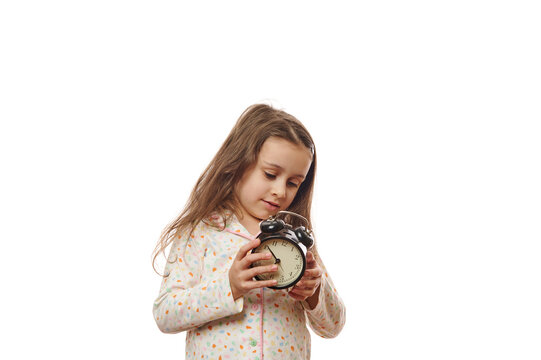 Caucasian Beautiful Sleepy Little Child Girl With Long Hair,wearing Trendy Soft Pajamas, Setting The Alarm Clock Before Going To Bed, Isolated On White Background With Copy Ad Space. It's The Bedtime