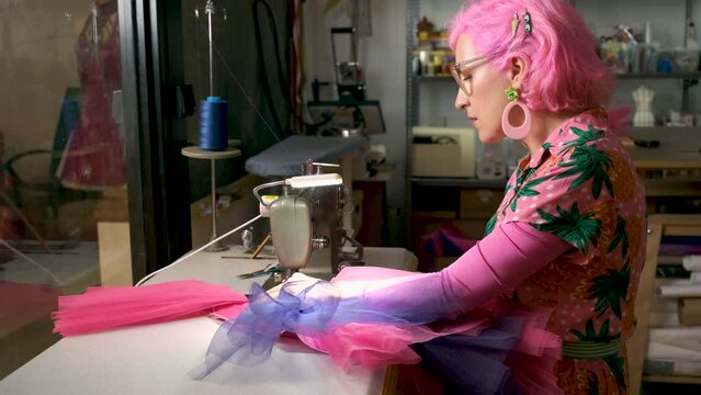 Dressmaker with pink hair and colorfull clothes smiling and looking at camera working on an industrial sewing machine in a sewing workshop.