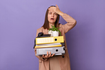 Horizontal shot of sad crying woman wearing beige jacket holding lot of documents folders isolated over lilac background., showing facepalm gesture, being fired.