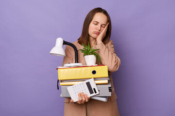 Portrait of exhausted overworked woman wearing beige jacket holding lot of documents folders isolated over lilac background, feels sleepy, leaning on her hand, standing with closed eyes.