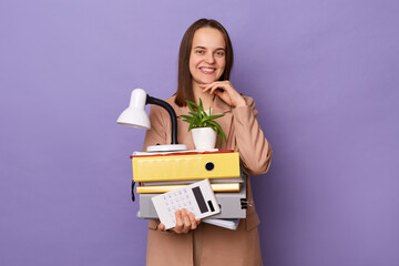 Indoor shot of smiling beautiful woman wearing beige jacket holding lot of documents folders,...