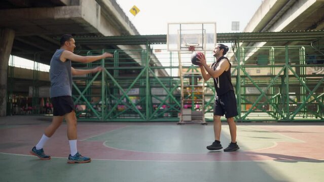 4K Asian man basketball player lifting his friend that fallen on outdoors court. Sportsman do sport training streetball at street court under highway in the city. Fair game sport competition concept.