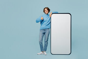 Full body fun young woman in knitted sweater near big huge blank screen mobile cell phone smartphone with workspace mockup area show thumb up like isolated on plain pastel light blue cyan background