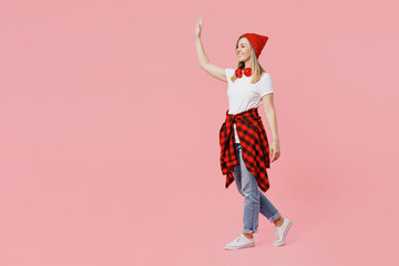 Full body smiling cheerful fun happy cool young woman wearing white t-shirt red hat walking going waving hand isolated on plain pastel light pink background studio portrait. People lifestyle concept.