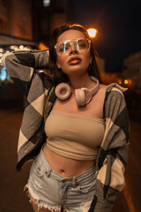 Fototapeta na wymiar beautiful woman with cool trendy glasses and pink headphones in fashion casual clothes with ripped jeans shorts, top and plaid shirt walks in the night city
