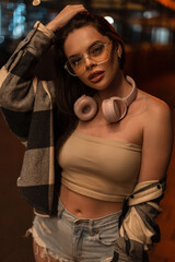 Fototapeta na wymiar Fashionable beautiful hipster woman with eyewear and headphones with top and plaid shirt with ripped jeans shorts walks in night city and fixed hair