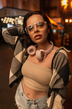 Cool young stylish hipster pretty girl with glasses and pink headphones in fashion street clothes with top, shirt and jeans shorts walking in the night city