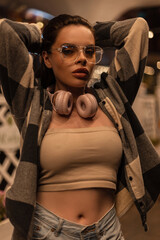 Fototapeta na wymiar Beautiful girl with fashion glasses and headphones in a stylish top with a plaid shirt stands on a night street
