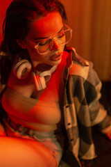 Stylish beautiful young woman with fashion glasses with headphones in a trendy top with a shirt and jeans shorts sits on the street with red light at night