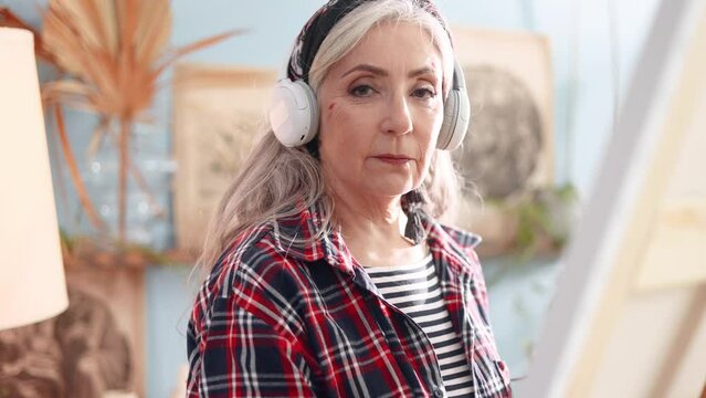 Senior woman artist listen music with headphones while painting picture on canvas creating artwork and looking at camera in creative workshop Mature grey haired lady enjoy leisure time with art hobby