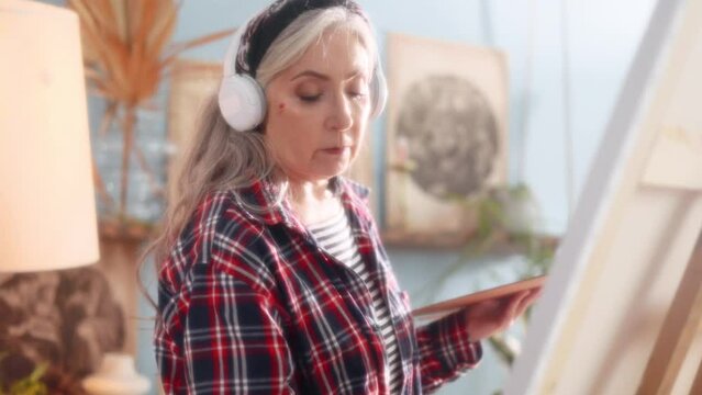 Dreamy soft of senior woman artist listen music with headphones while painting picture on canvas creating artwork in sunny creative workshop Mature grey haired lady enjoy leisure time with art hobby