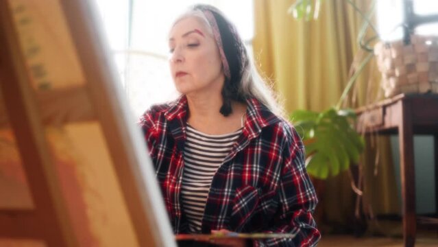 Dreamy soft shot senior woman artist painting picture on canvas creating artwork sits on floor in the light sunny creative workshop Stylish mature grey haired lady enjoying leisure time with art hobby