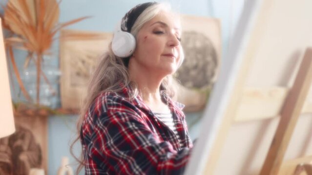 Dreamy soft of senior woman artist listen music with headphones while painting picture on canvas creating artwork in sunny creative workshop Mature grey haired lady enjoy leisure time with art hobby