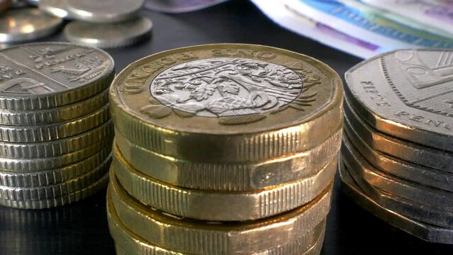 Closeup slow zoom shot of fingers adding a pile of 50 pence pieces next to British pound and other coins and sterling notes.