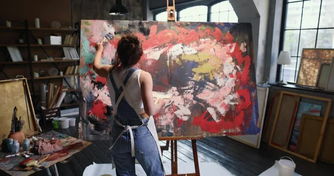 Beautiful Caucasian artist creates abstract masterpiece with paint brush. Woman artist paints colorful abstract painting with brush on huge canvas. Back view of artist works on emotional piece of art.