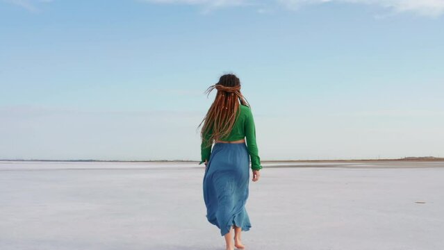 Girl dressed in boho style walking barefoot on surface of lake. Back view. Unrecognizable young woman with dreadlocks is going on pond shore in windy summer day. Promenade on river bank