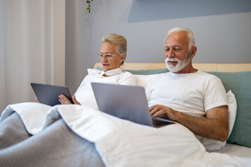 Senior couple lying on bed with tablet and laptop