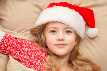 a happy blonde girl in a Santa Claus hat and New Year's pajamas is lying in bed