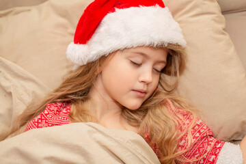 a beautiful blonde girl in a Santa Claus hat and New Year's pajamas is sleeping in bed