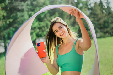 Excited fit beautiful brunette girl in green sportswear holds yoga mat, phone prepares for exercise outdoors toothy smiles looks at camera against green trees at park. Sporty hispanic woman at workout