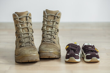 old worn military boots and children's sports shoes on wooden floor. Concept of military father and...