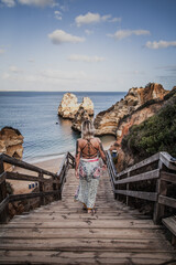a tattooed woman in a dress walking down wooden stairs leading to a beautiful cliff sand beach strand in Portugal