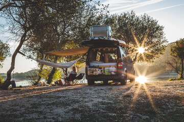 A sunset camping scene in the wild with a camper van in between trees on a passing by river. - obrazy, fototapety, plakaty