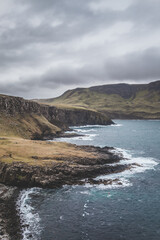 Fototapeta na wymiar waves crushing onto the ruff rocky coastal line of the Isle of Skye in Scotland, cloudy grey sky and yellow brown green grass surrounded by cliffs