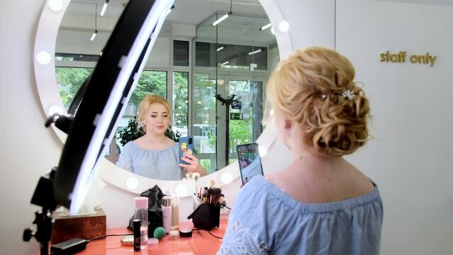 Young pretty woman makes photos near lit mirror using smartphone camera. Blonde lady enjoys modern hairstyle and make-up from famous masters in beauty salon