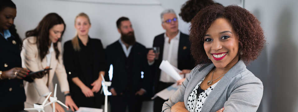 Close-up latin businesswoman posing at camera with smile. Teamwork working at desk office on background. Horizontal banner or header.