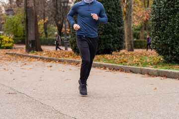 Unrecognizable sportsman running on asphalt path covered with dry leaves during fitness workout in autumn park