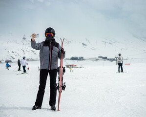 Fototapeta na wymiar Girl dressed in a ski suit and helmet with balaclava stands with ski against the backdrop of snow-covered mountain ski slope, cloudy sky, village below and take selfie on smartphone. Winter. Sport