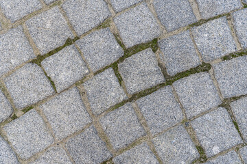 Natural stone background and texture in gray color close up. Pavement paved with square and uneven stones. Paving street with pattern. Between crushed cobblestones there are earth sand and grass.