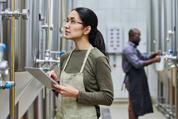 Young female engineer using digital tablet to check pressure in barrels with brewed beer