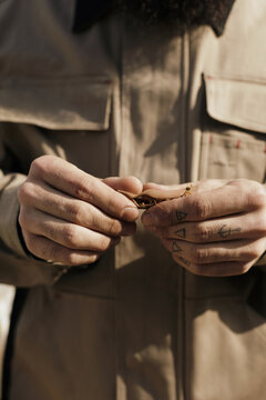 Close-up shot of unrecognizable man with small tattoos on fingers in workwear rolling up tobacco cigarette