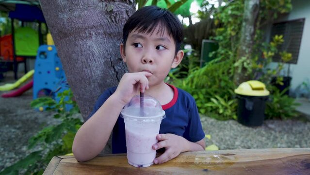 4K, An Asian boy, sitting in park with playground, Sitting drinking water, Pipo Yogurt smoothie, looking tired from playing toys playground, Wears blue shirt, with red stripe on collar.