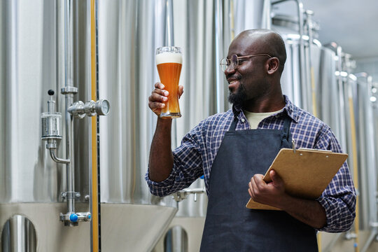 Smiling African American worker holding glass of fresh beer to check the quality after brewing while standing in workshop with tanks