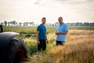 Zwei Landwirte stehen an einem reifen Getreidefeld, Vorderrad eines Treckers ist zu erkennen.