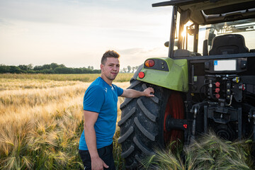 Junger Landwirt steht an einen Hinterrad eines Treckers im reifen Getreidefeld.