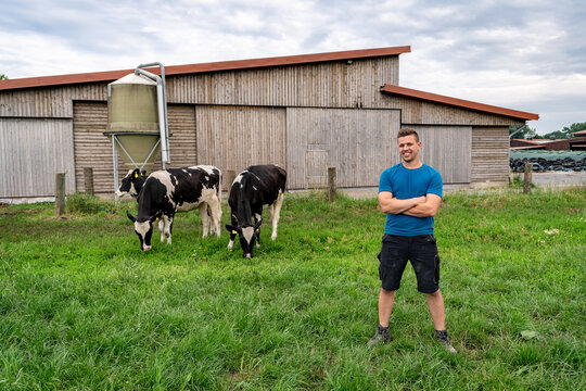 Junglandwirt steht stolz auf einer Wiese mit Rindern, im Hintergrund ein Rinderstall.