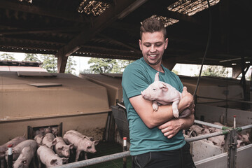 Junger Landwirt freut sich über gesunde Ferkel, er hält eines auf den Arm vor einer Ferkelbucht die mit Ferkeln gefüllt ist. © Countrypixel