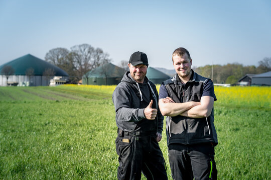 Hofnachfolger und Vater stehen in einem Getreidefeld und sind zufrieden, Biogasanlage im Hintergrund.
