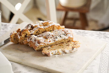 Traditional german christmas pastry stollen on a marble cutting board, coffee mug, butter plate on the table cloth and scandy style kitchen chair in the back