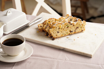 Traditional german christmas pastry stollen on a marble cutting board, coffee mug, butter plate on the table cloth and scandy style kitchen chair in the back